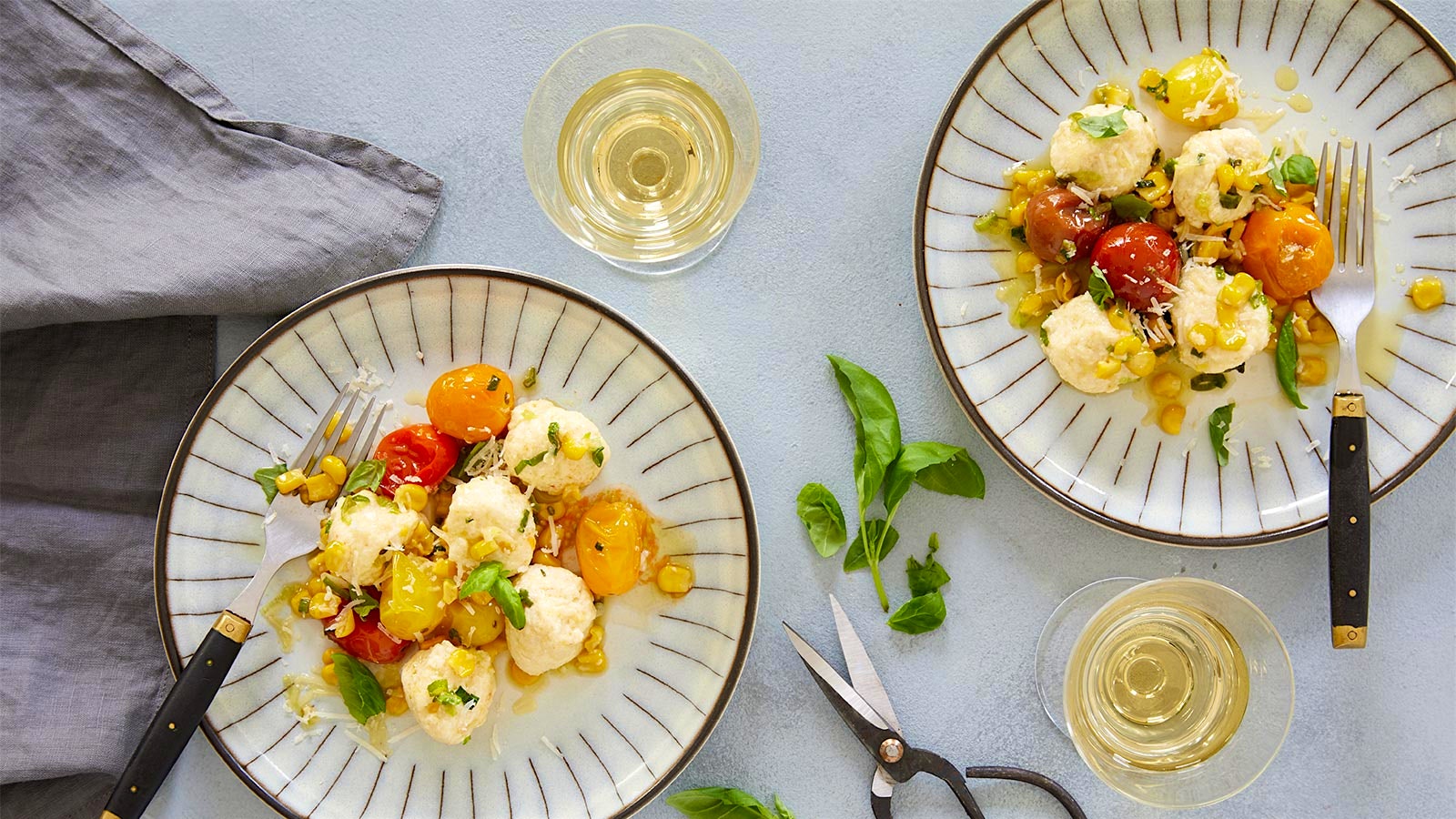 Two plates of gnudi dumplings mixed with tomatoes and corn are decorated with fresh basil and placed next to two white wine glasses